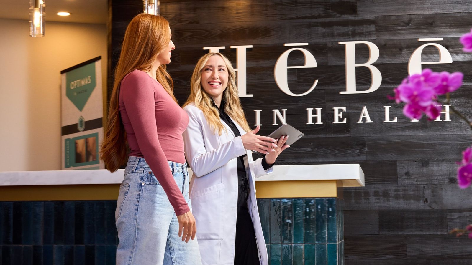 Nicole Smith, MSPA, in a white coat reviewing a treatment plan on a tablet with a patient at the HēBē Skin Health front desk